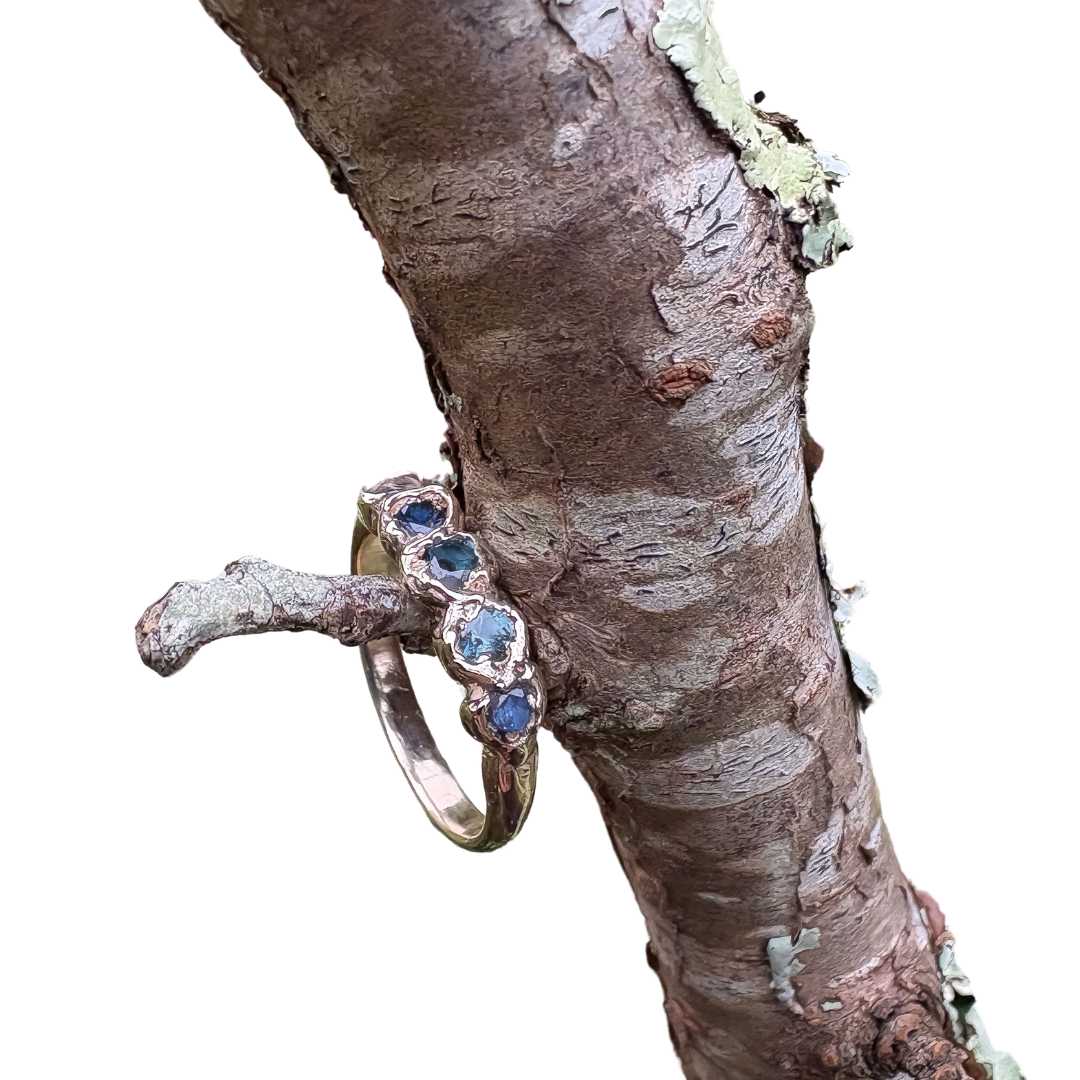 A gold ring with a row of blue gemstones, shown resting on a textured tree branch against a white background.