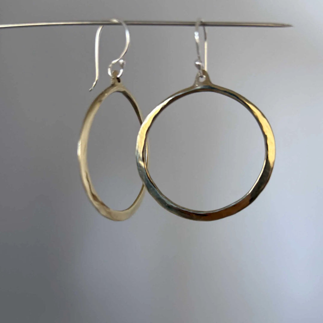 A pair of hammered bronze hoop earrings hanging from silver fishhook wires against a soft gray gradient background.