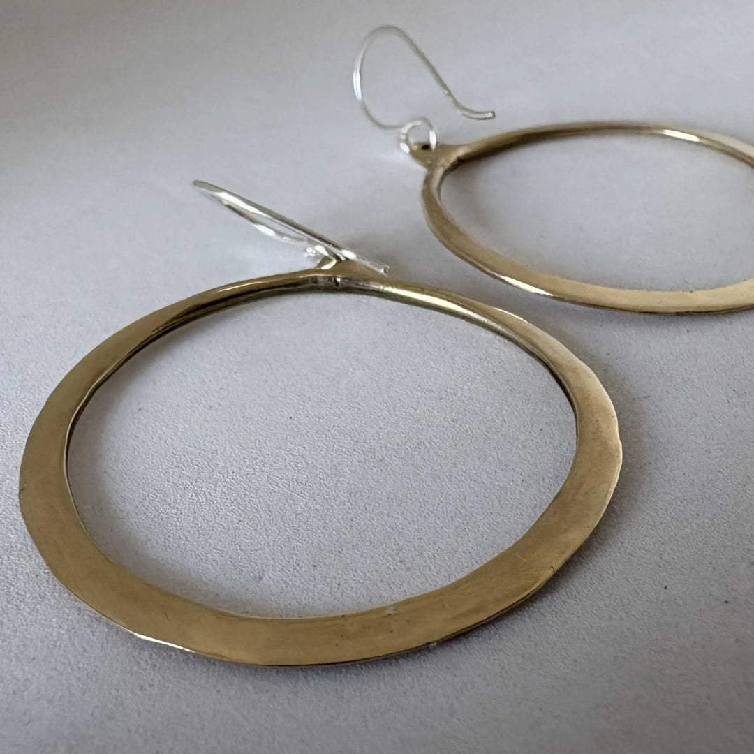 Large hammered gold-tone hoop earrings with silver fishhook wires, displayed on a light gray textured surface.