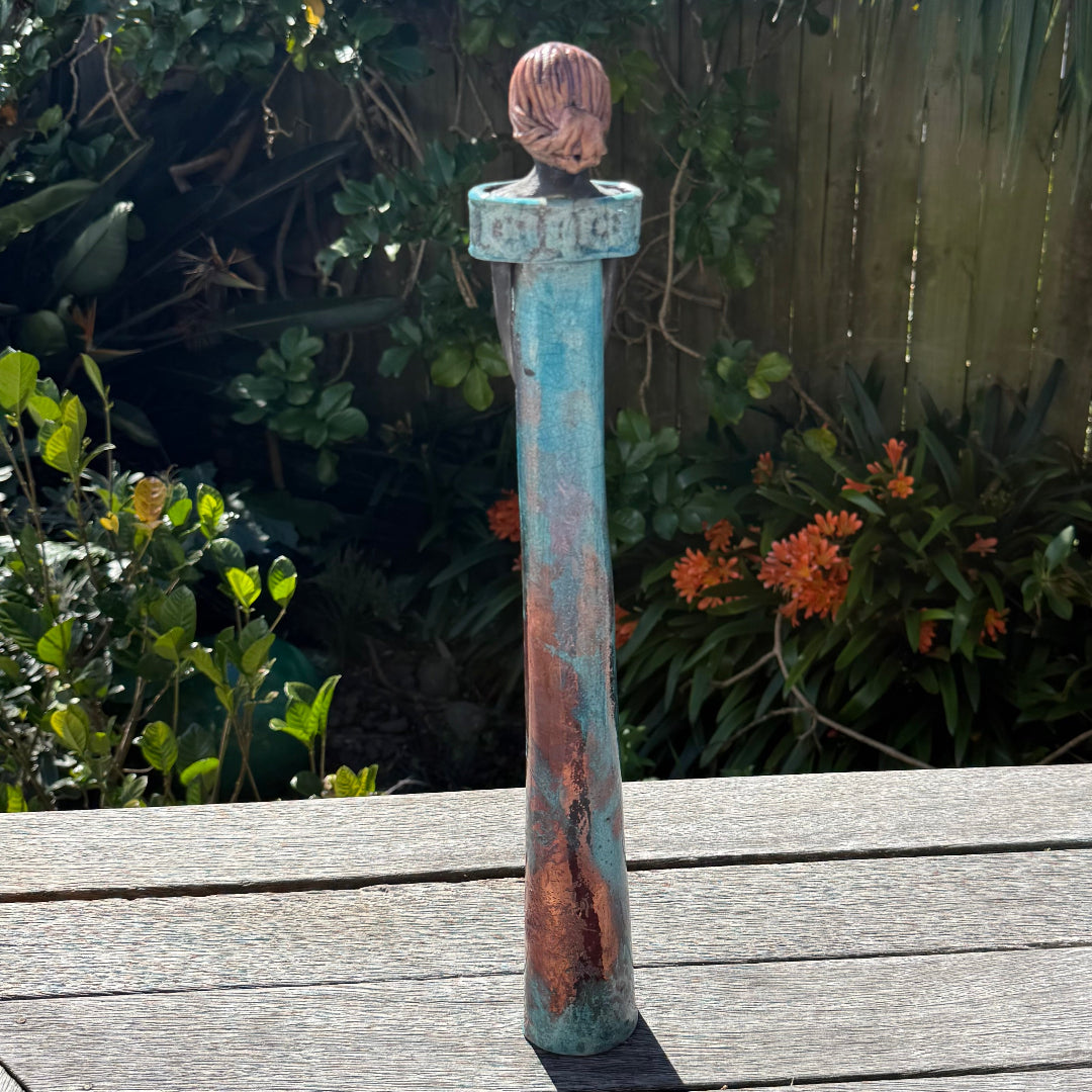 Back view of a tall lady ceramic figure with a distressed turquoise-blue and rust-orange glaze, photographed outdoors on a wooden table with garden foliage and a fence in the background.
