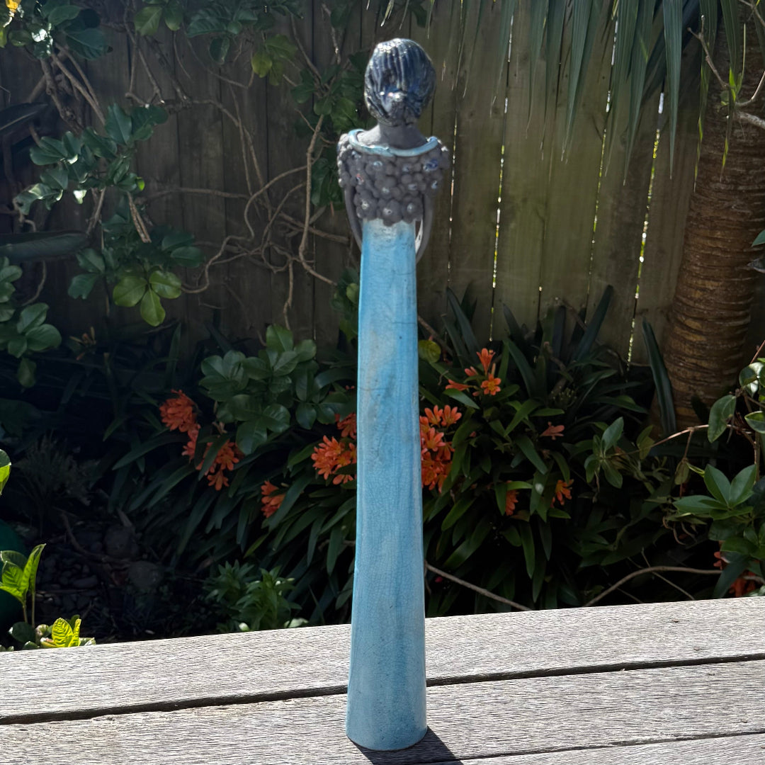 Back view of a tall lady ceramic figure with an aqua glazed dress & dark floral bodice, photographed outdoors on a wooden table with garden foliage and a fence in the background.