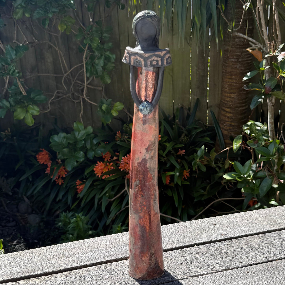 A tall lady ceramic figure with a shiny copper glazed dress with patterned collar holding a bouquet, photographed outdoors on a wooden table with garden foliage and a fence in the background.