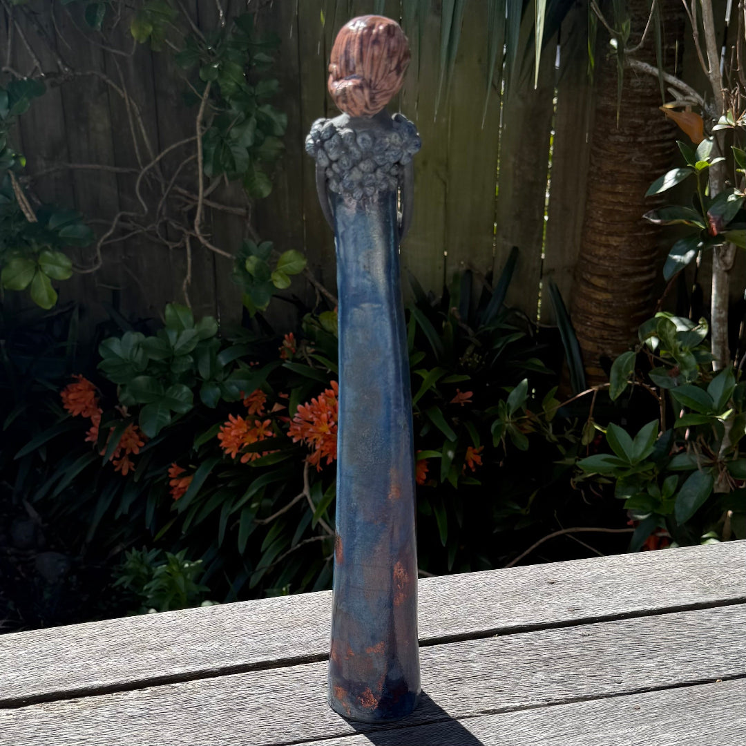 Back view of a tall lady ceramic figure with a distressed turquoise-blue and rust-orange glaze, photographed outdoors on a wooden table with garden foliage and a fence in the background.