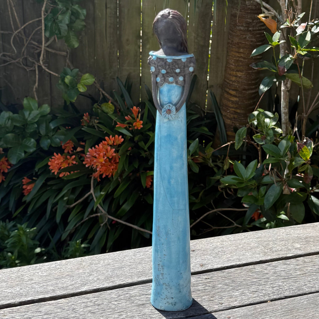 A tall lady ceramic figure with an aqua glazed dress & dark floral bodice, holding an aqua flower, photographed outdoors on a wooden table with garden foliage and a fence in the background.