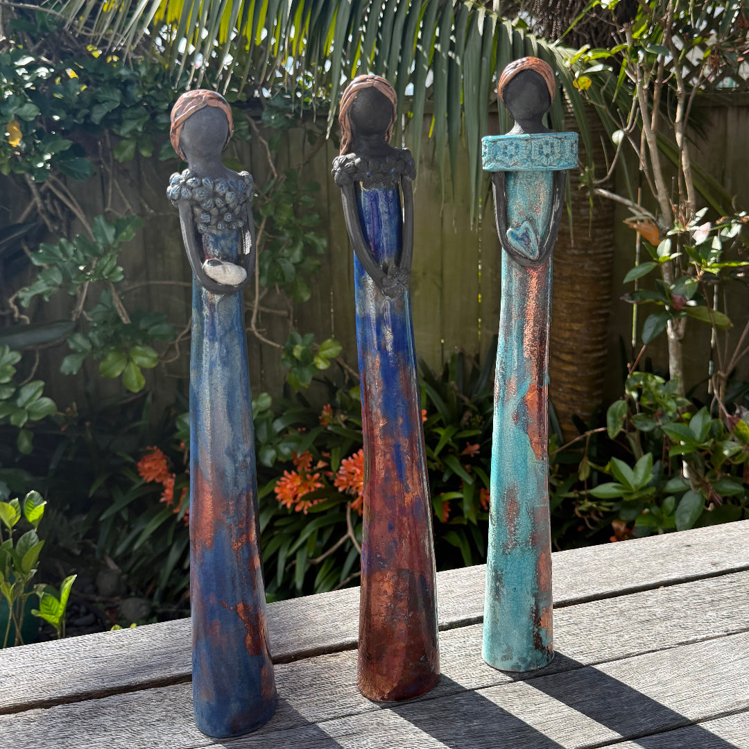 Three tall ceramic ladies figurines with black faces and elongated bodies stand on a weathered wooden deck outdoors, with tropical plants and a blue sky in the background.