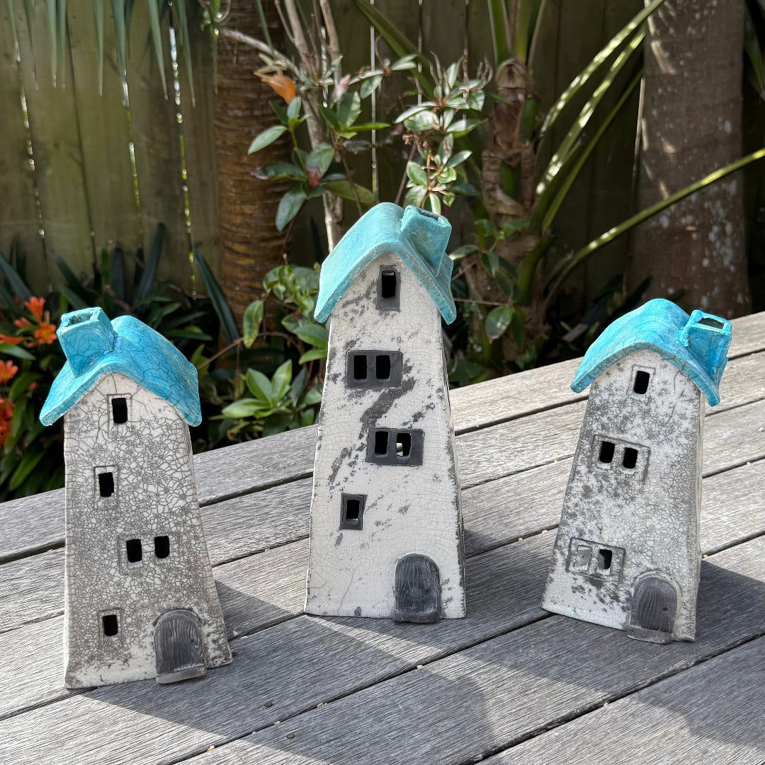3 ceramic house-shaped tealight holders with distressed white bodies, window cutouts, and coloured roofs, photographed outdoors on a weathered wooden surface with garden plants in the background.