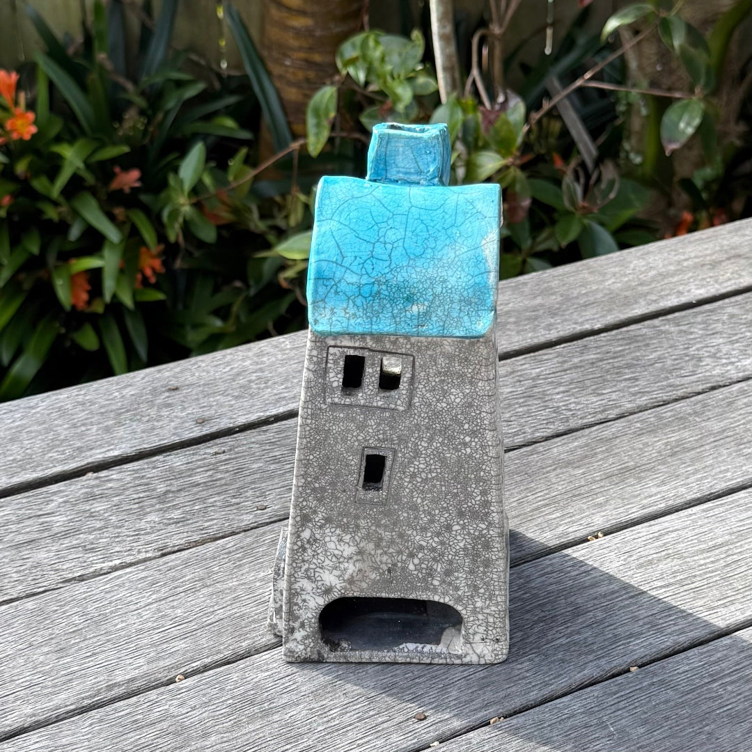 A ceramic house-shaped tealight holder with a distressed white body, dark gray window cutouts, and an aqua roof, photographed outdoors on a weathered wooden surface with garden plants in the background.