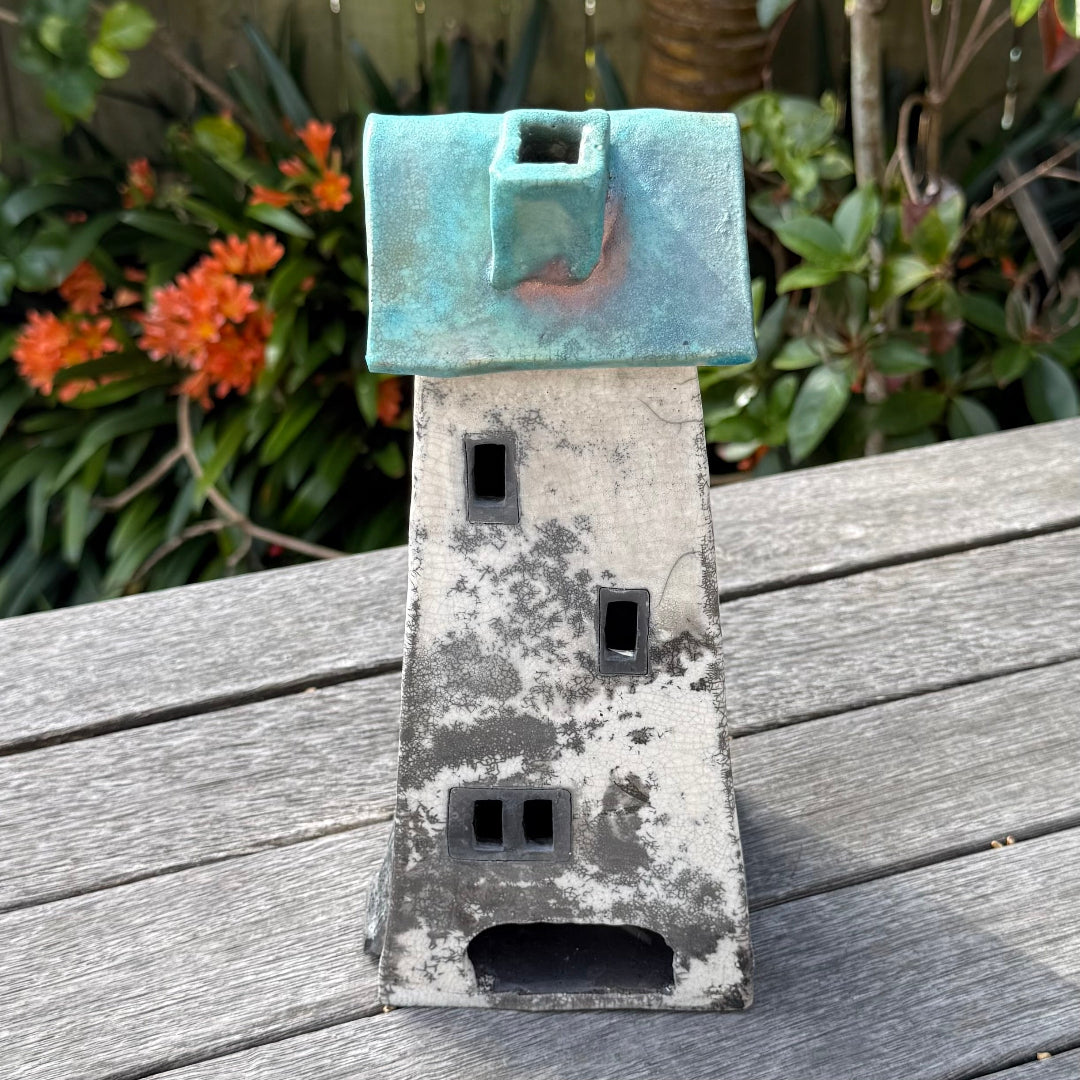 A ceramic house-shaped tealight holder with a distressed white body, dark gray window cutouts, and a turquoise roof, photographed outdoors on a weathered wooden surface with garden plants in the background.