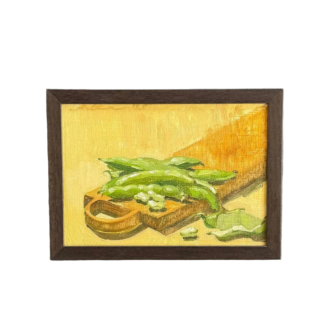 A framed still life oil painting depicting green beans on a wooden cutting board, mounted on a white wall.