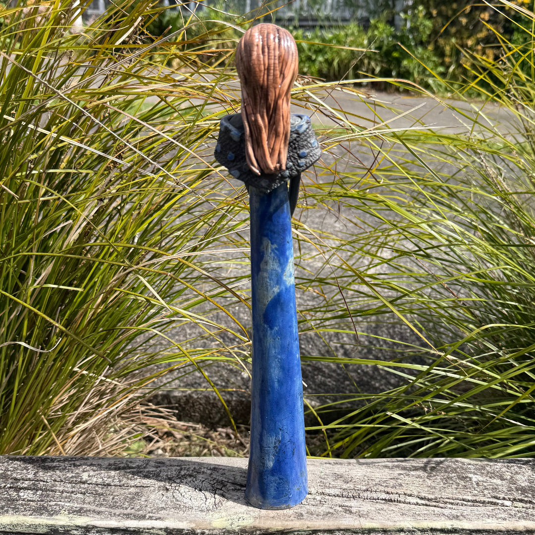 Back view of a decorative statue of a woman with long blonde hair and a blue patina body, standing on a weathered wooden deck with garden foliage in the background.