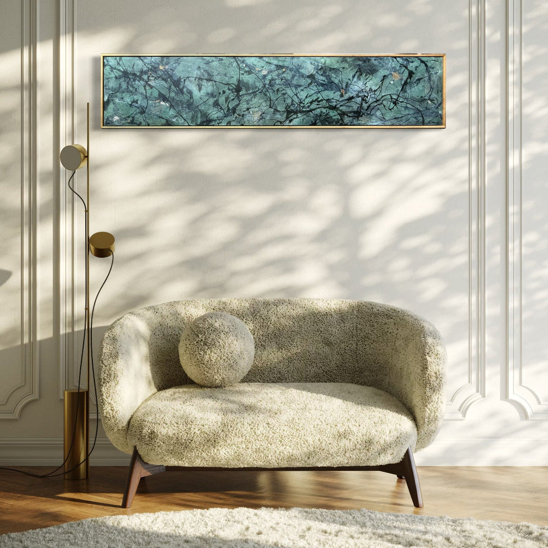 A long horizontal framed wall art piece with a teal and dark blue marbled pattern hangs on a cream paneled wall above a textured beige armchair in a sunlit room.