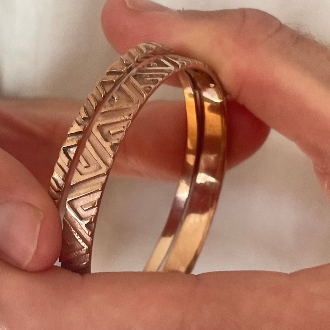 Copper & silver bangles with geometric pattern held between fingers against a neutral background