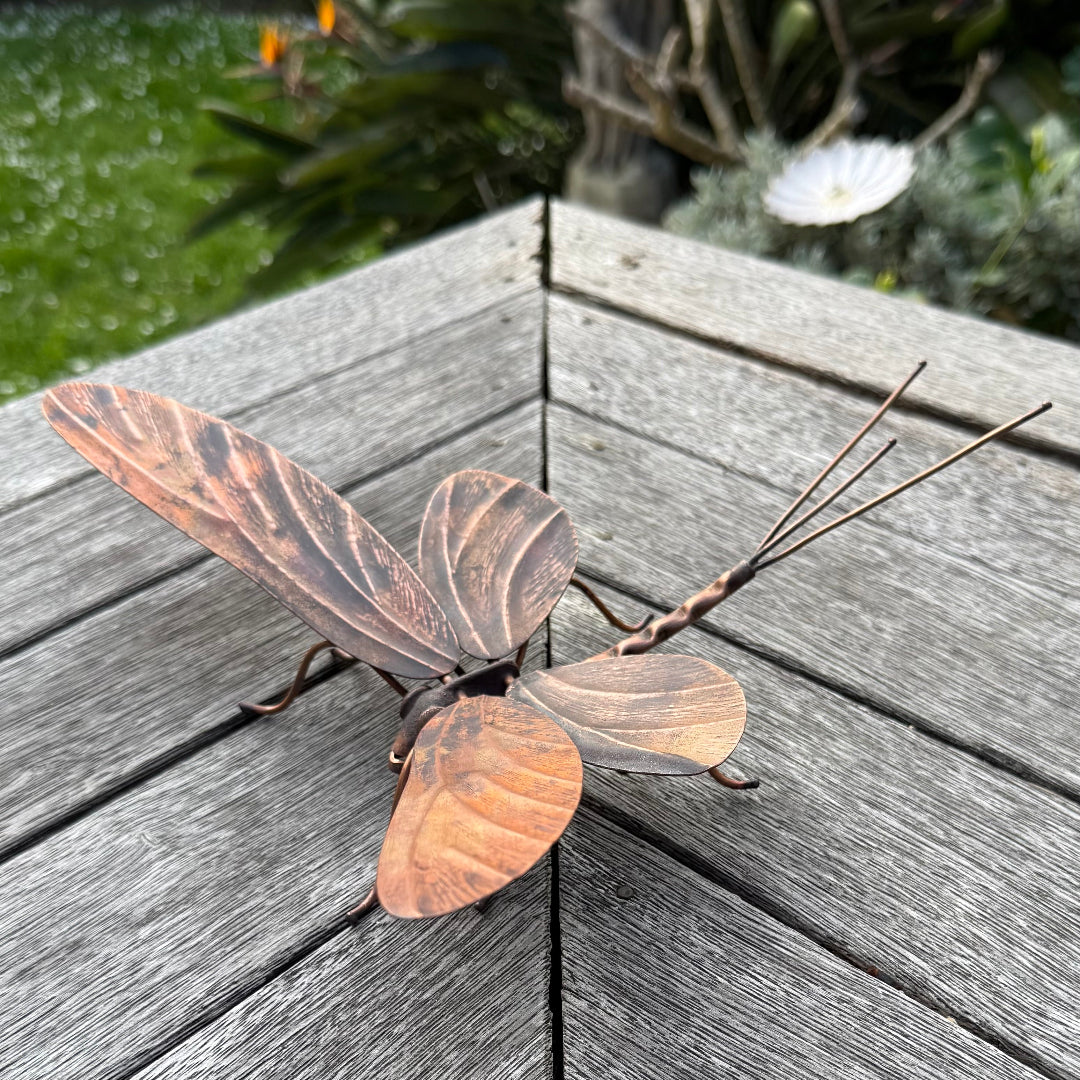 Copper Mayfly sculpture on a wooden surface with a garden background