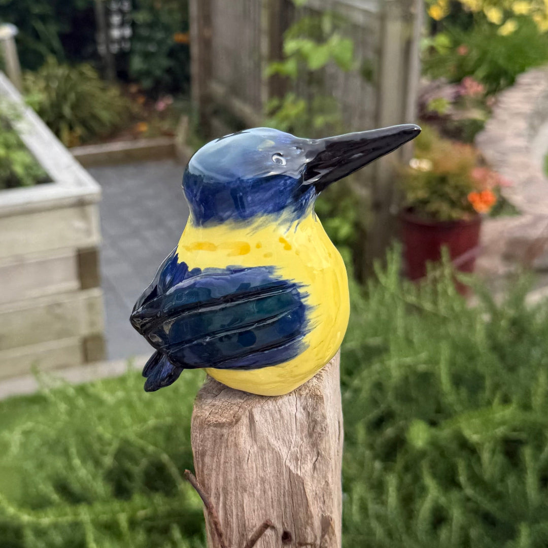Ceramic Kingfisher bird with blue and yellow colors on a wooden post in a garden setting.