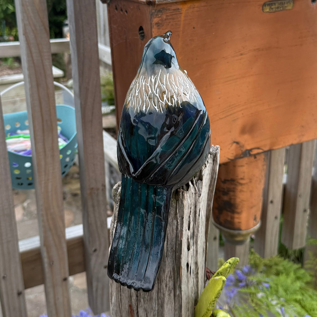 Back view of a ceramic Tui bird sculpture on a wooden post with a garden background