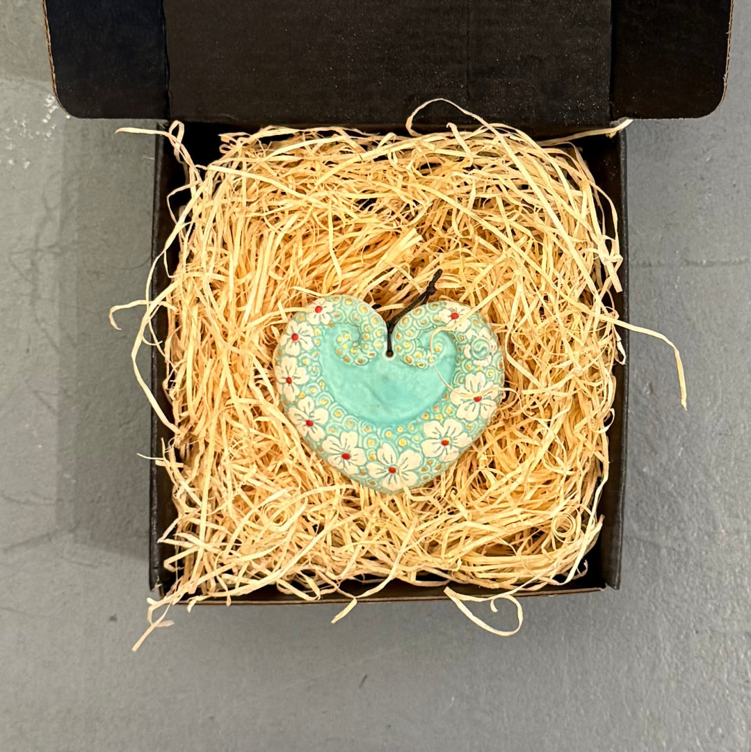 Decorative ceramic heart-shaped wall hanging in a nest of straw inside an open black box on a grey surface.