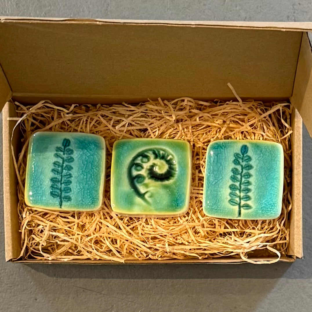 Three square ceramic tiles with botanical designs in a box lined with straw