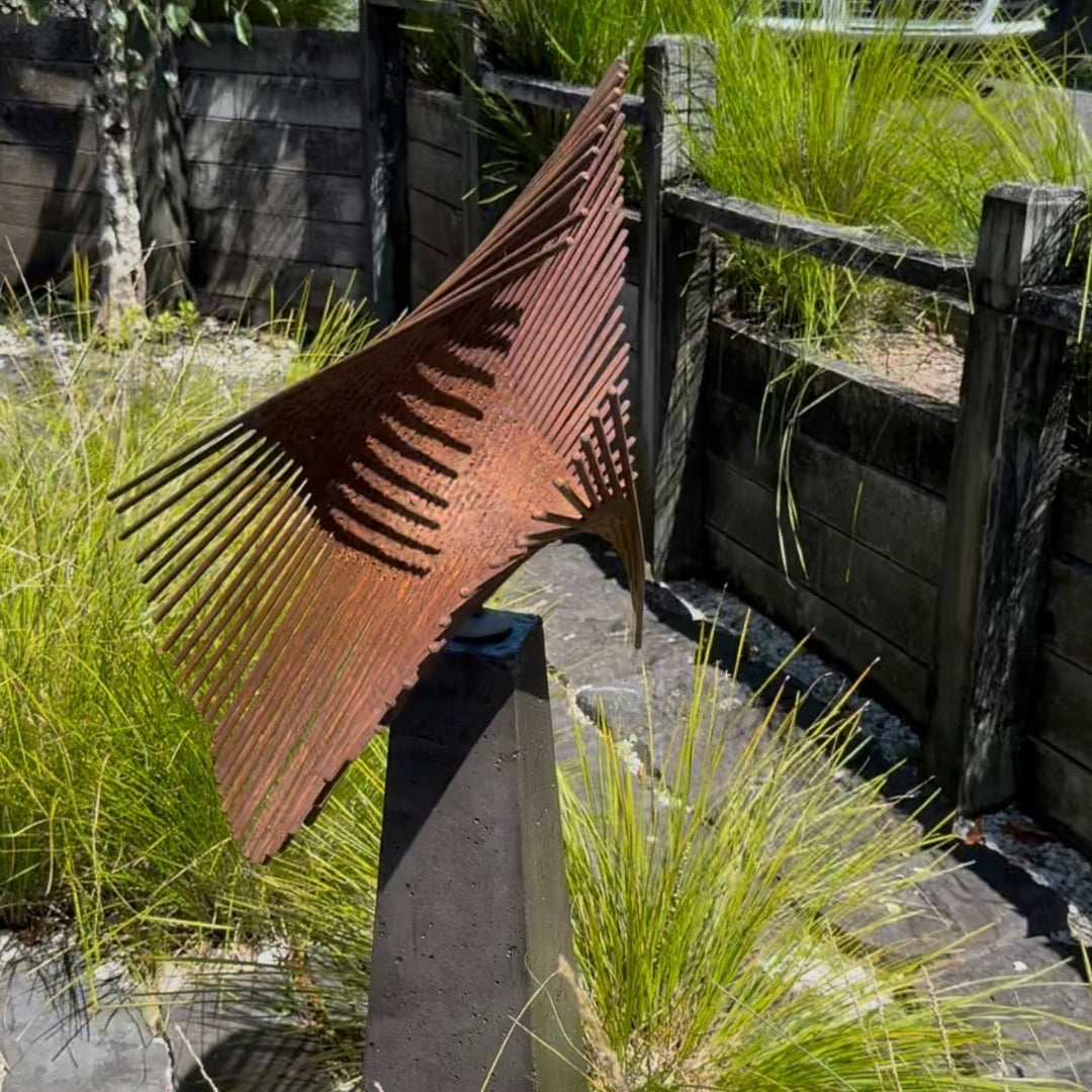 Rust-colored metal sculpture on a stone pedestal in a garden setting with grass and wooden barriers.