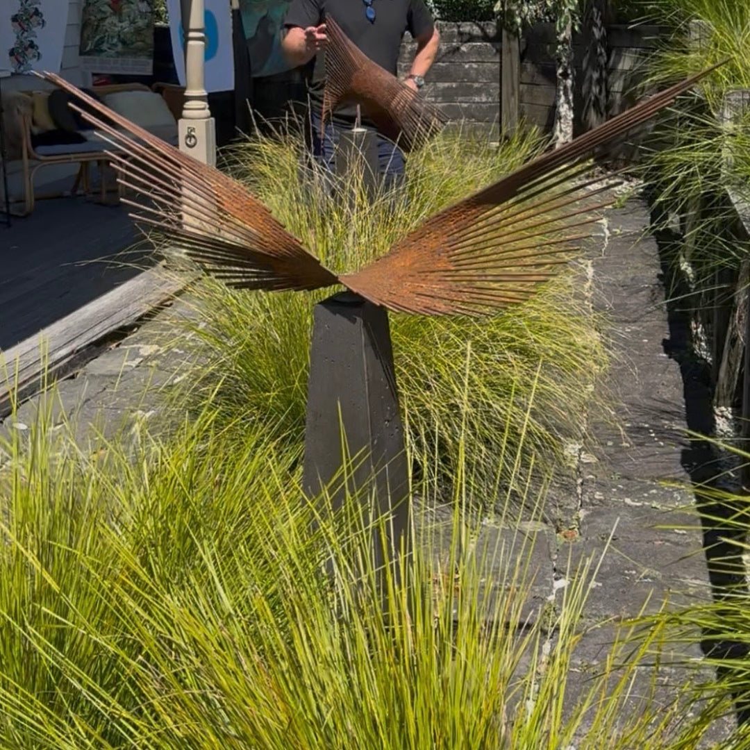 Decorative metal sculpture resembling a bird in flight surrounded by greenery on a stone path.