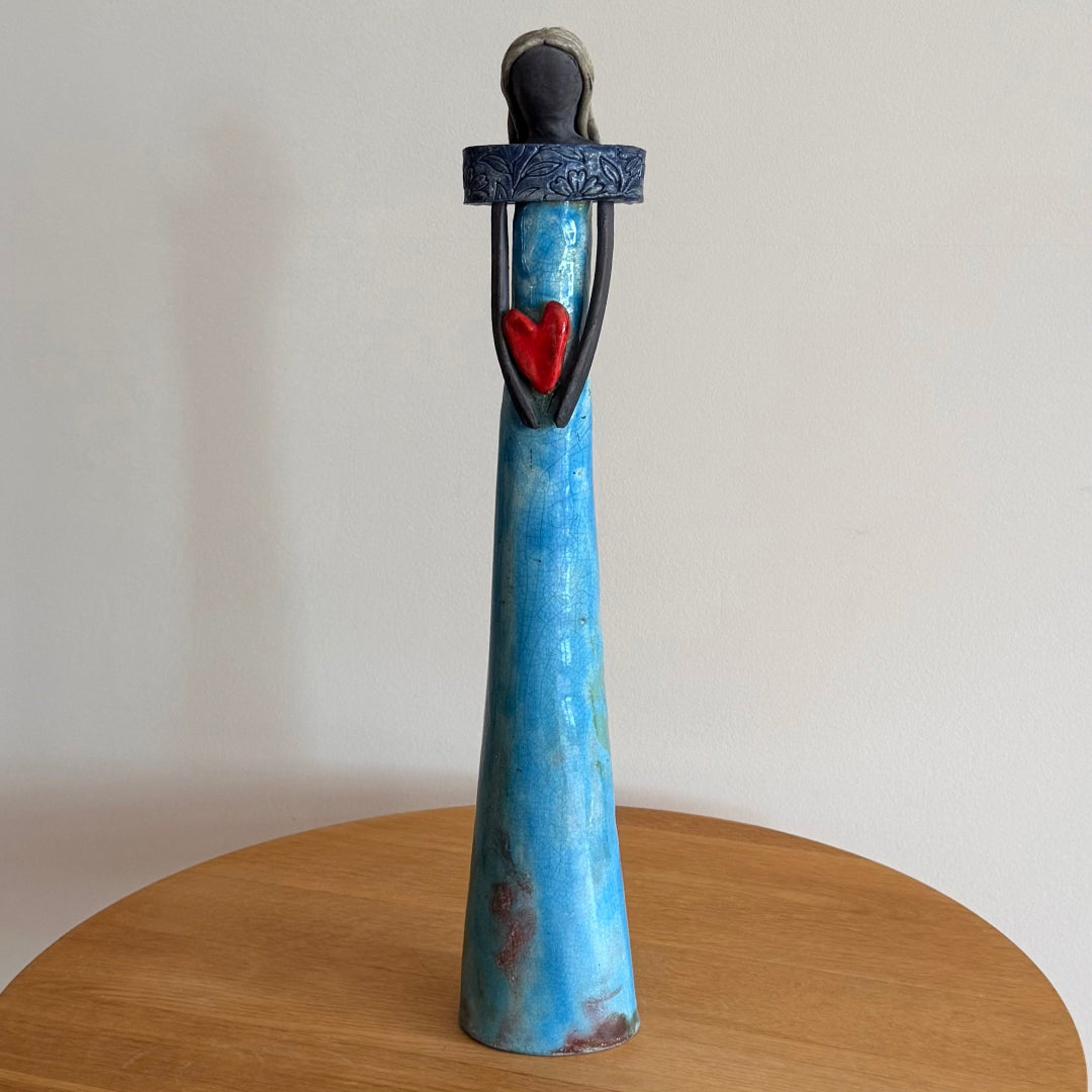 Ceramic figurine with blue dress, red heart, and navy collar on a wooden surface.