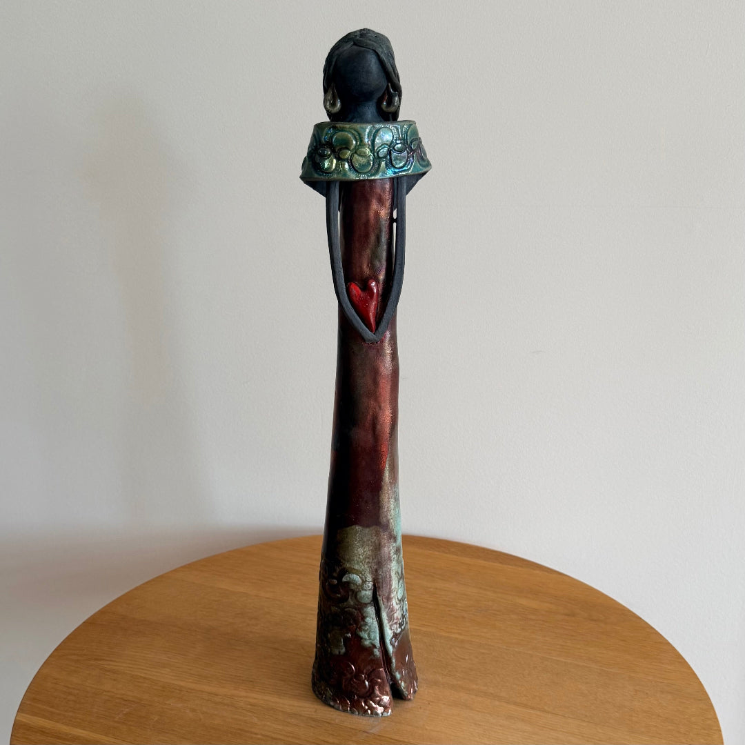Tall ceramic woman with green earrings & collar holding a red heart on a wooden surface