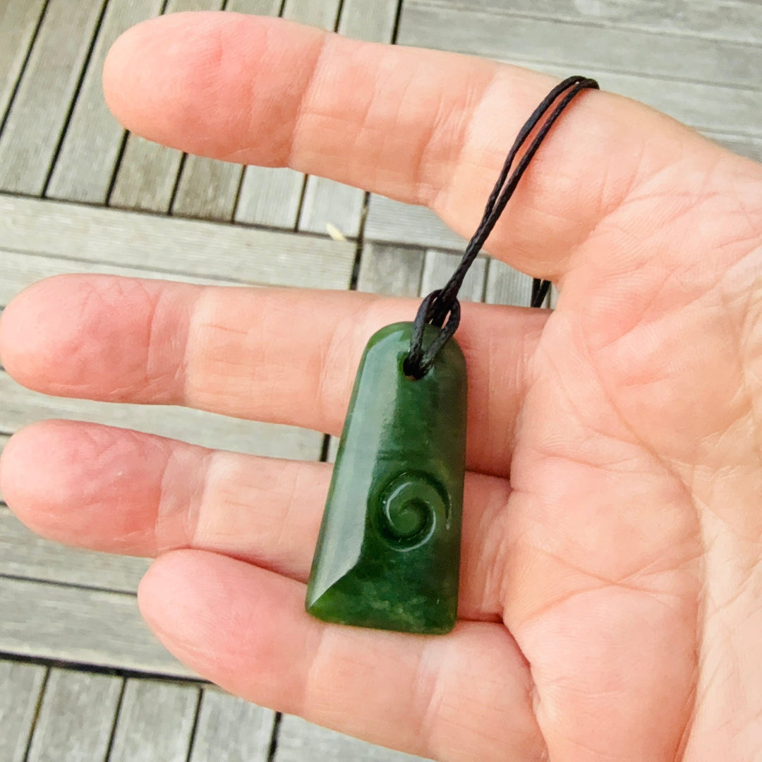 Hand holding a green nz jade toki pendant with a black cord against a wooden backgroundki