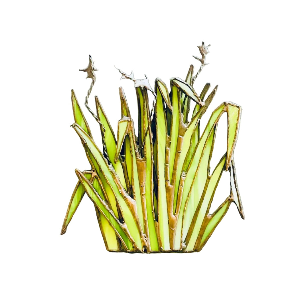 A decorative stained glass wall-mounted sculpture shaped like a clump of green and yellow Harakeke or flax, against a white wall.