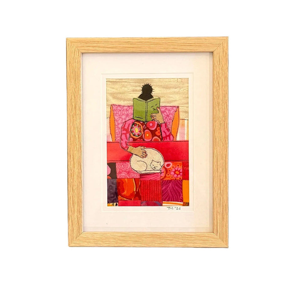 Framed handstitched textile artwork of a woman in pink reading a book with a cat on her lap on a white background.