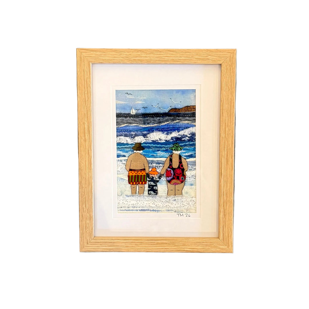Framed textile artwork of three people on a beach with a wooden frame against a white wall.
