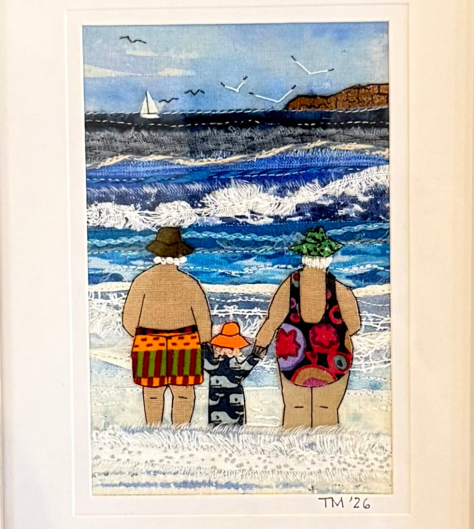 Textile artwork of three people in swimsuits on a beach with ocean and cliffs in the background.