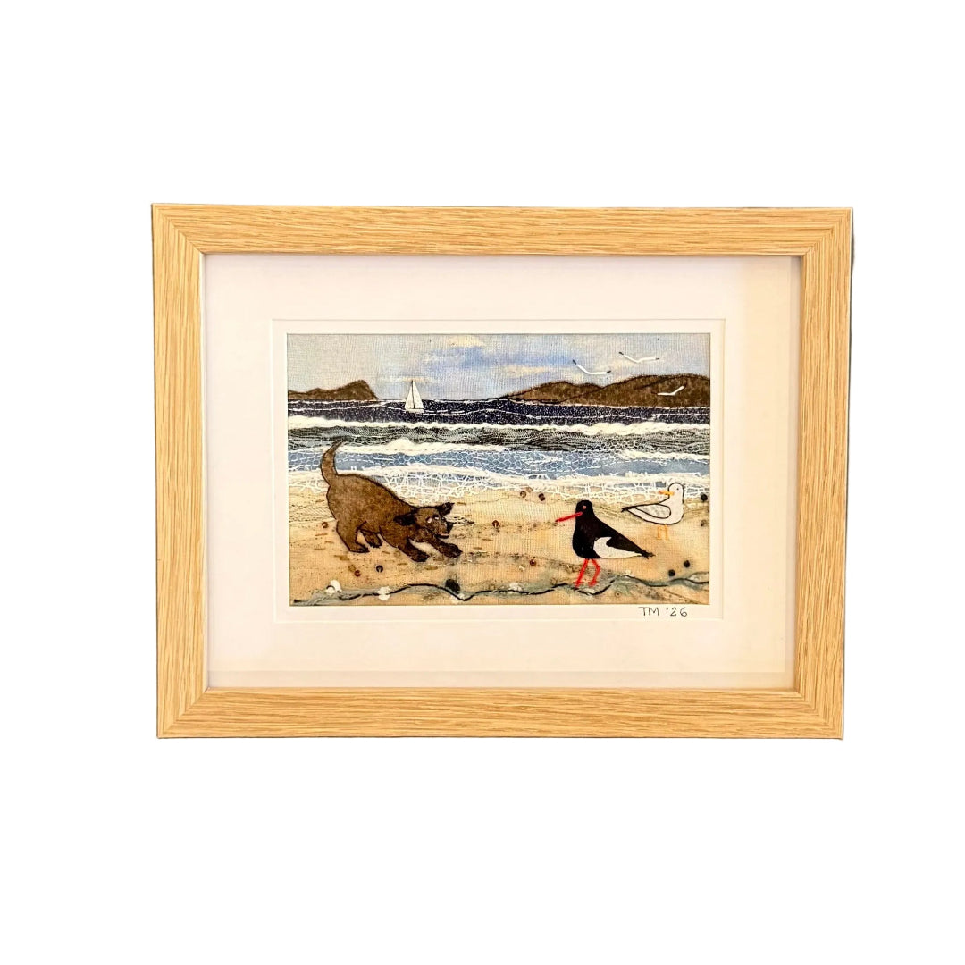 Framed textile artwork of a dog and oystercatcher on a beach against a white wall