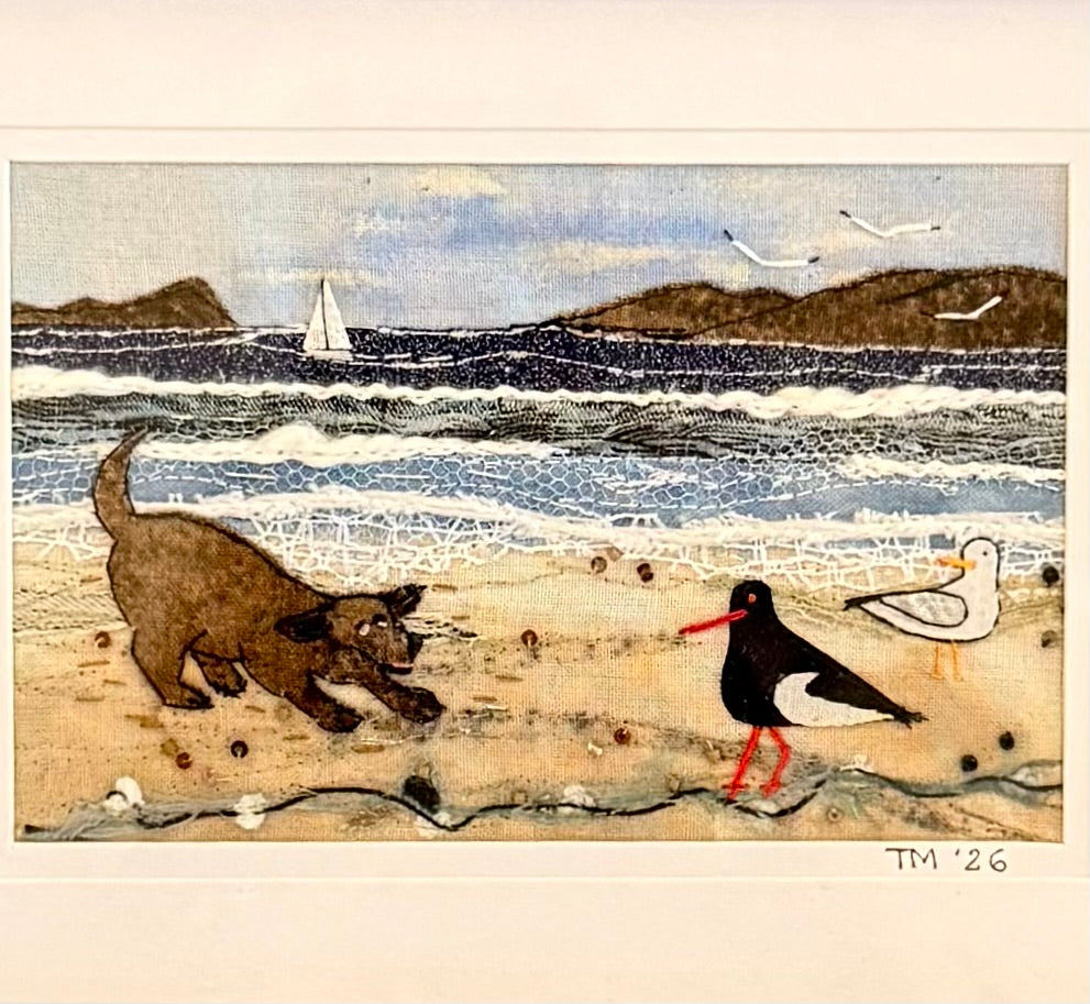 Textile artwork of a dog chasing birds on a beach with a sailboat in the background.