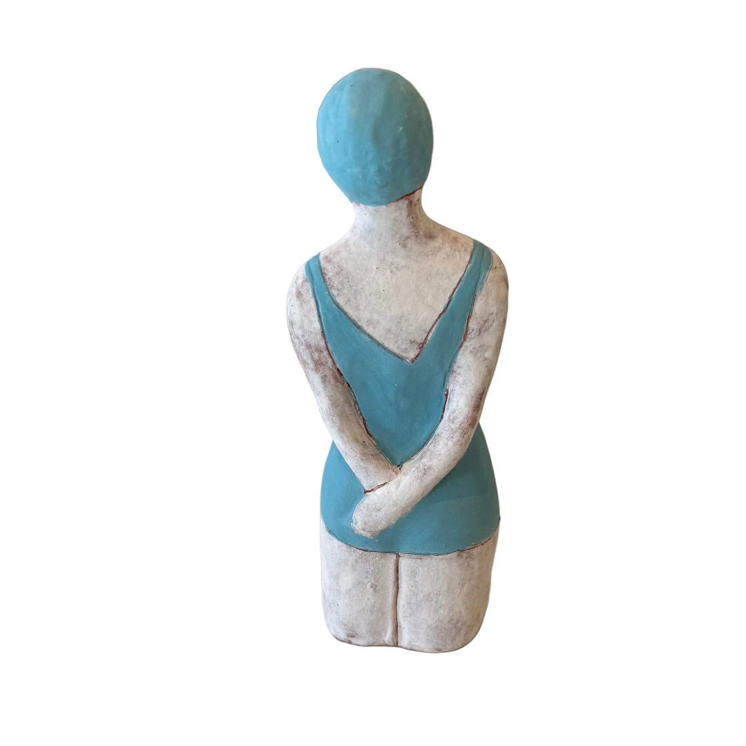 Back view of a ceramic sculpture of a Swimmer in blue swimsuit & cap with hands behind her back on a white background