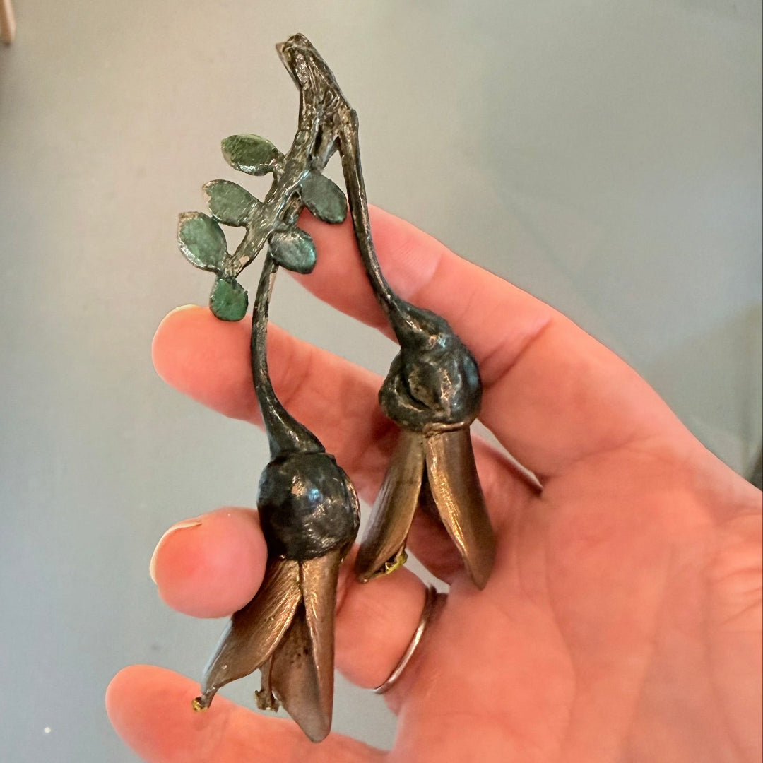 Hand holding a pair of miniature painted bronze Kowhai flowers with visible seed pods, placed on a plain background.