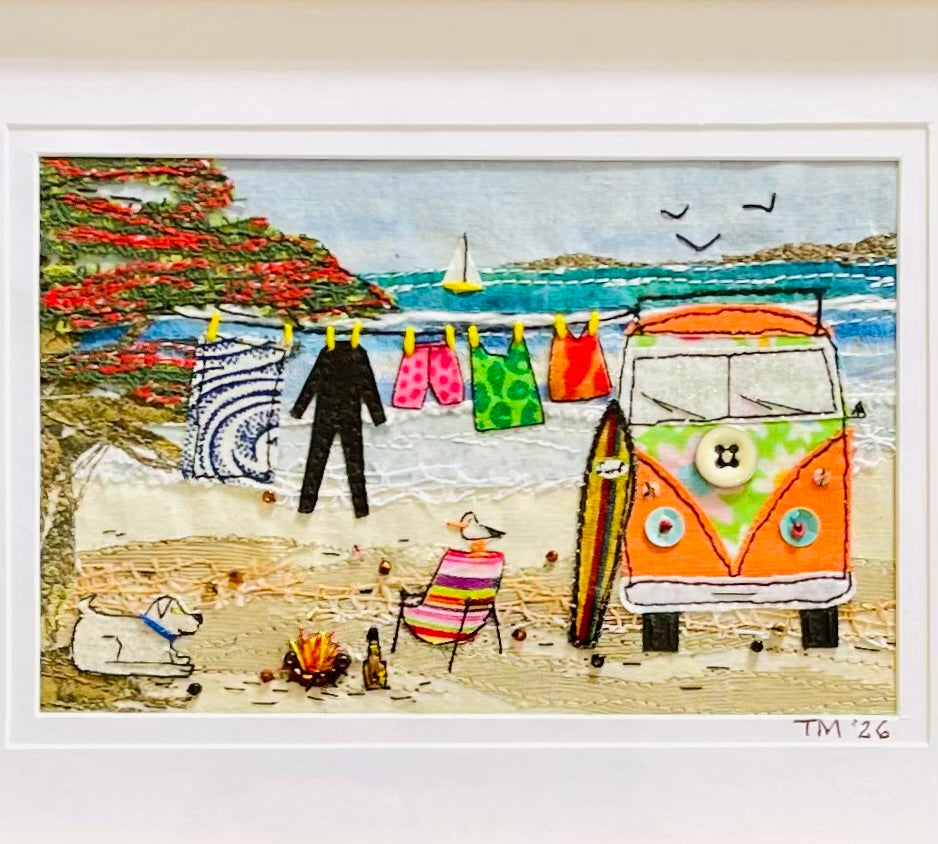 Framed beach scene textile artwork showing an orange and green retro van on a sandy shore, colorful laundry hanging on a line, a surfboard, a striped lounge chair, a small dog, and the ocean in the background.