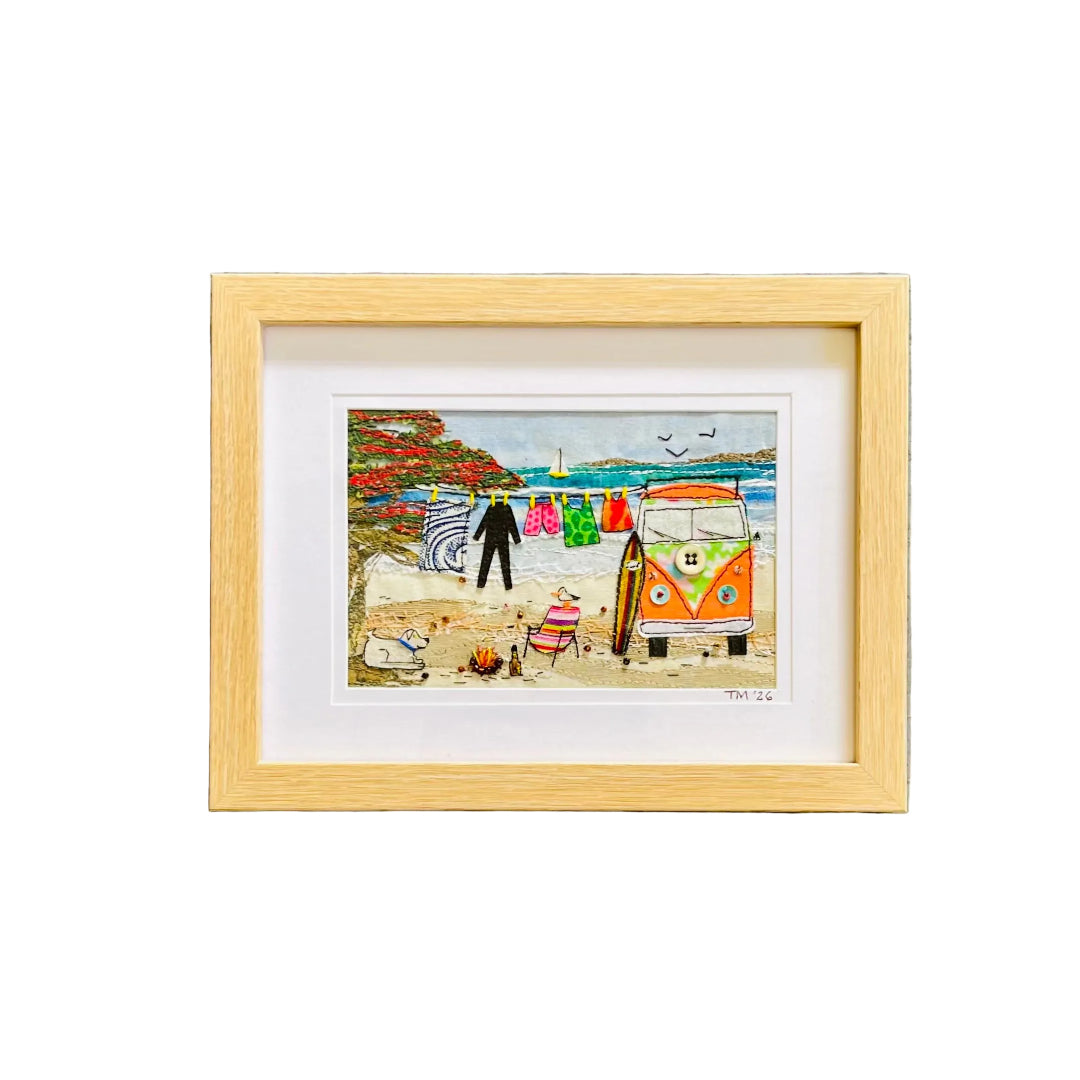 Framed textile art depicting a colorful beach scene with a vintage orange and white VW bus, a striped towel, and beach umbrellas, set in a light wood frame against a white wall.