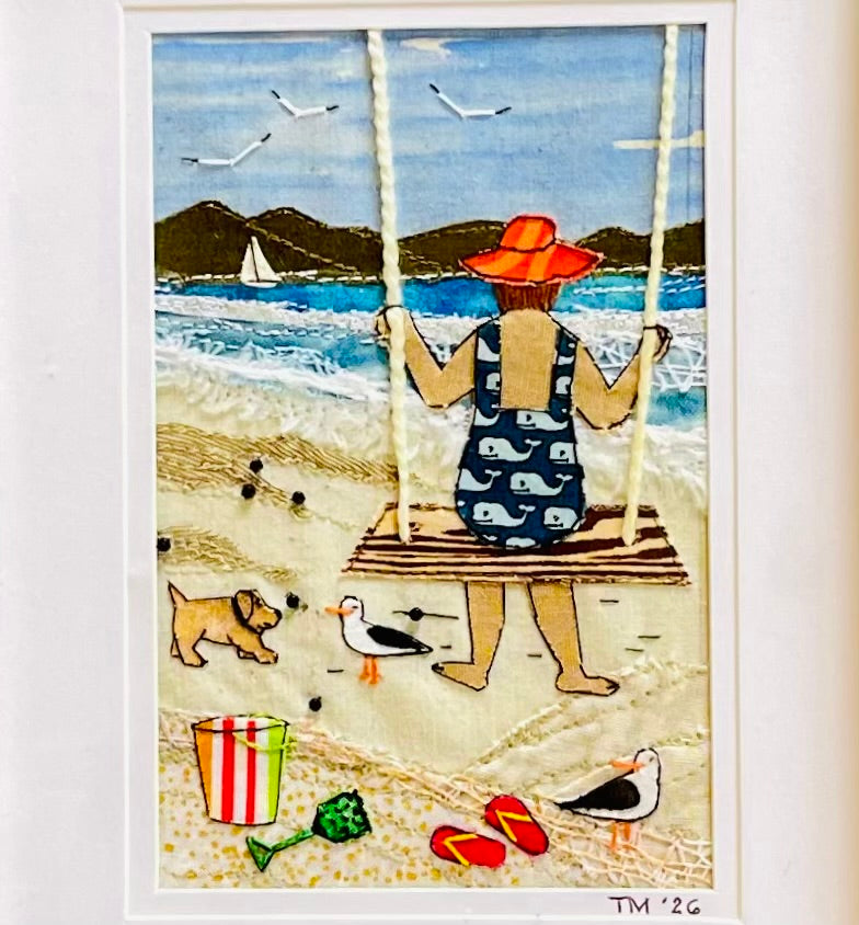Framed textile art depicting a woman sitting on a beach swing, viewed from behind, with dogs, seagulls, and the sea in the background. The print is matted and housed in a light natural wood frame, mounted on a white wall.