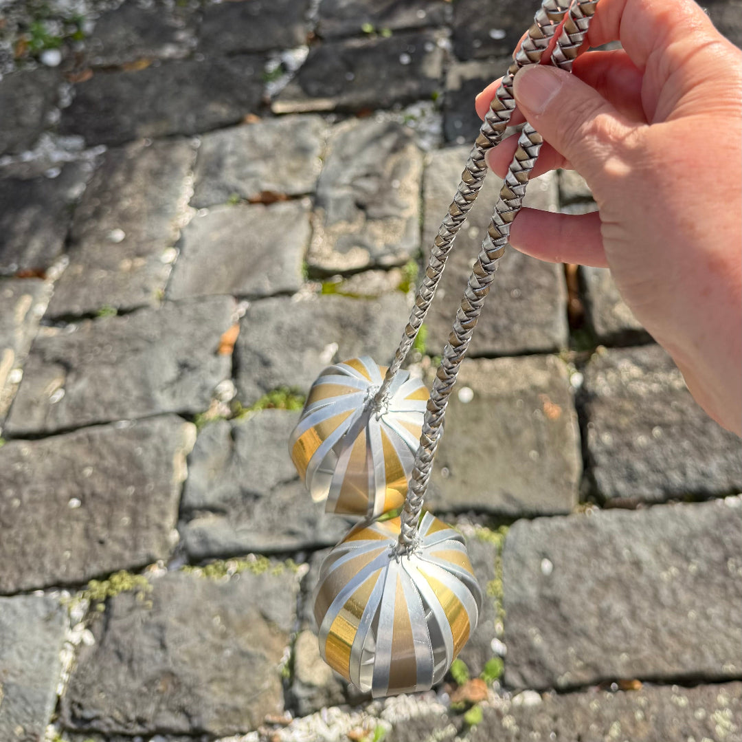 Hand holding a pair of silver & gold aluminium poi with leaf-like top on a stone pavement background