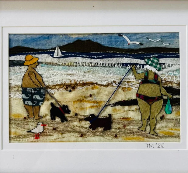 Textile art of two people with dogs on a beach scene.