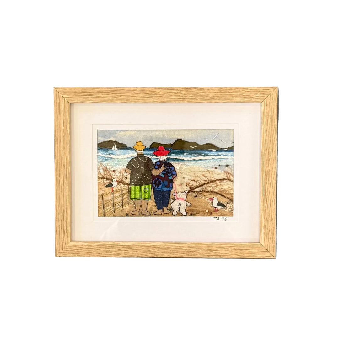 Framed textile artwork of a couple by the beach with a dog on a white wall.