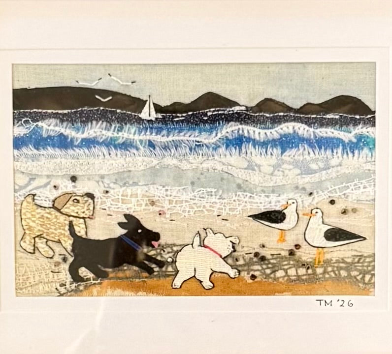 Textile artwork of dogs and seagulls by a coastal scene with mountains and a sailboat.