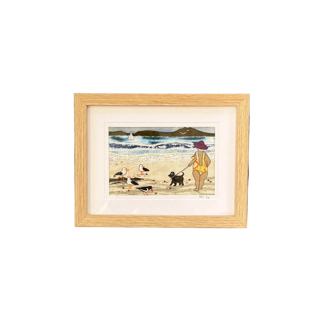 Framed textile artwork of a person with a dog on a beach, hanging on a white wall.
