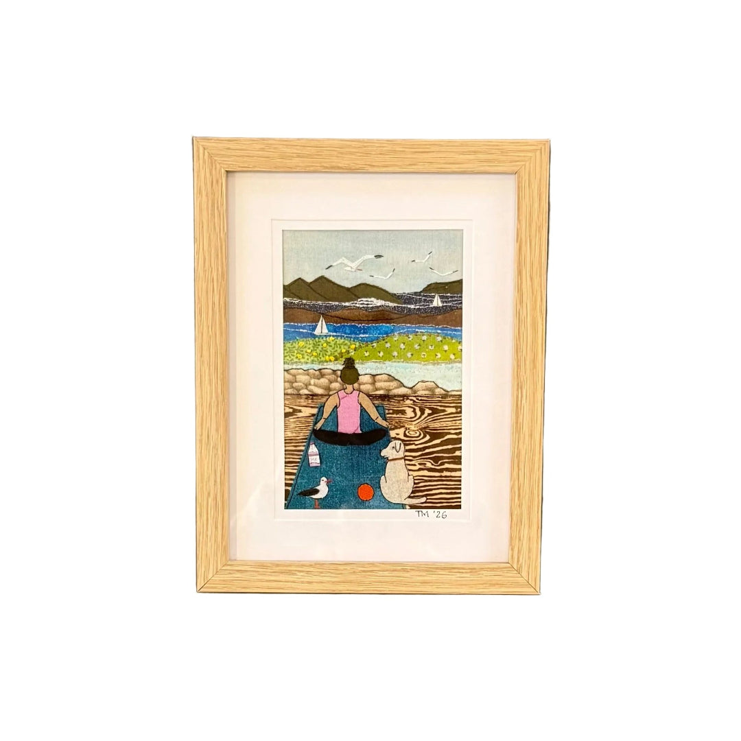 Framed textile artwork of a woman with a dog on a dock with mountains and sailboats in the background.