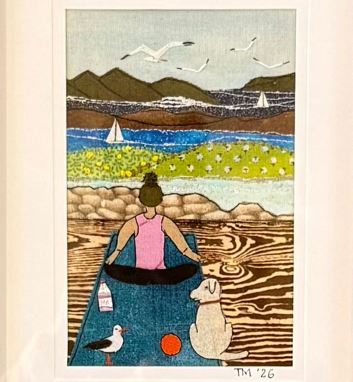 Textile artwork of a woman sitting on a yoga mat by a beach with mountains and seagulls.