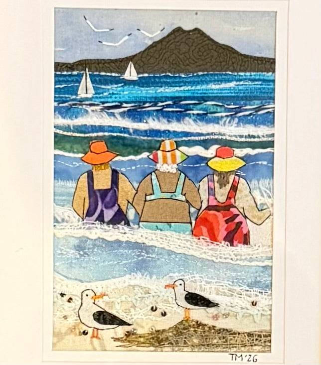 Textile artwork of three people in swimsuits and hats on a beach with seagulls and sailboats in the background.