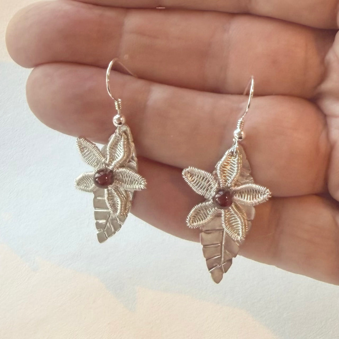 Floral silver wire-wrapped earrings held between fingers against a neutral background
