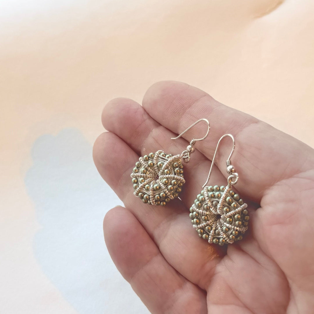 Hand holding a pair of silver & green pearl kina shaped earrings on a beige background