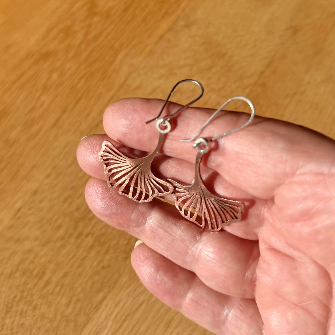 Hand holding a pair of silver ginkgo leaf earrings against a wooden background