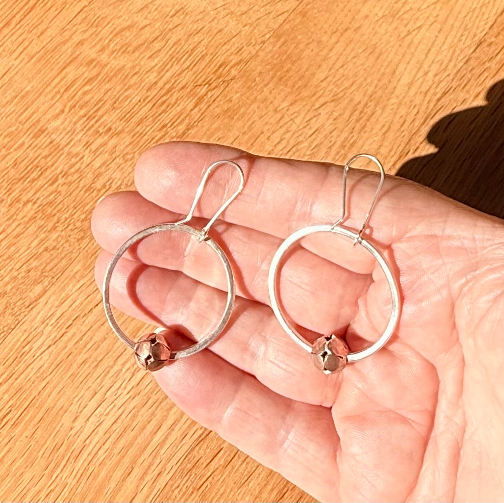 Hand holding two silver hoop earrings with small round charms on a wooden surface