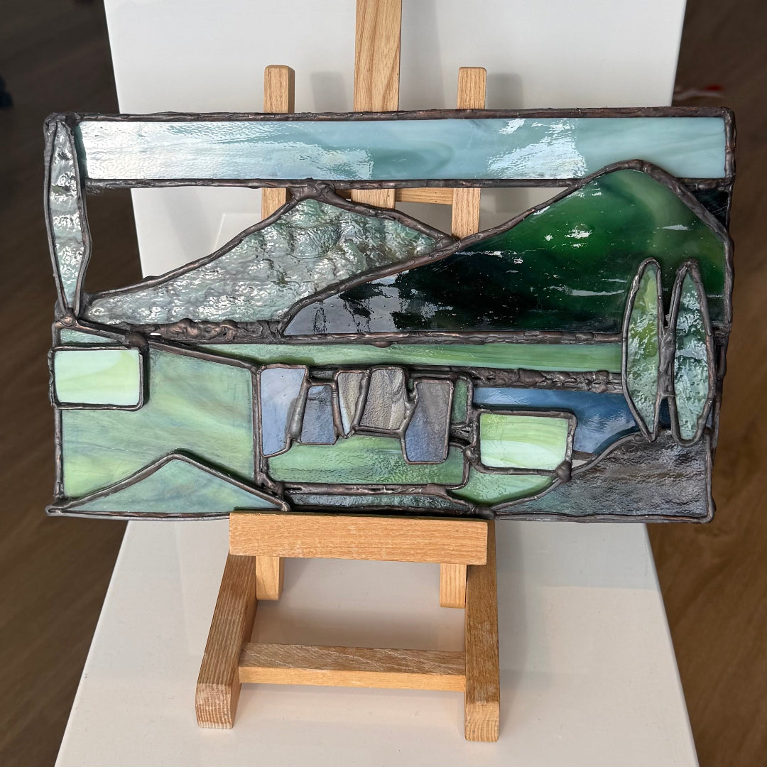 A stained glass panel depicting a mountain landscape with green hills, blue water, and gray mountains, displayed on a small wooden easel stand.